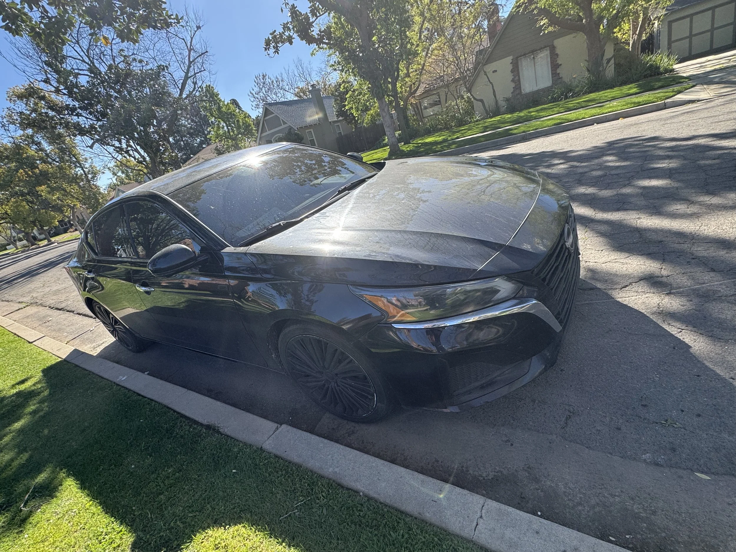 A black sedan parked on a residential street under a partially cloudy sky, with houses, trees, and greenery in the background.