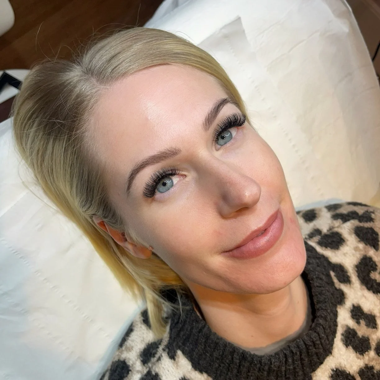 Close-up of a woman with blonde hair, blue eyes, and applied makeup, lying on a white pillow, wearing a black and white leopard print sweater.