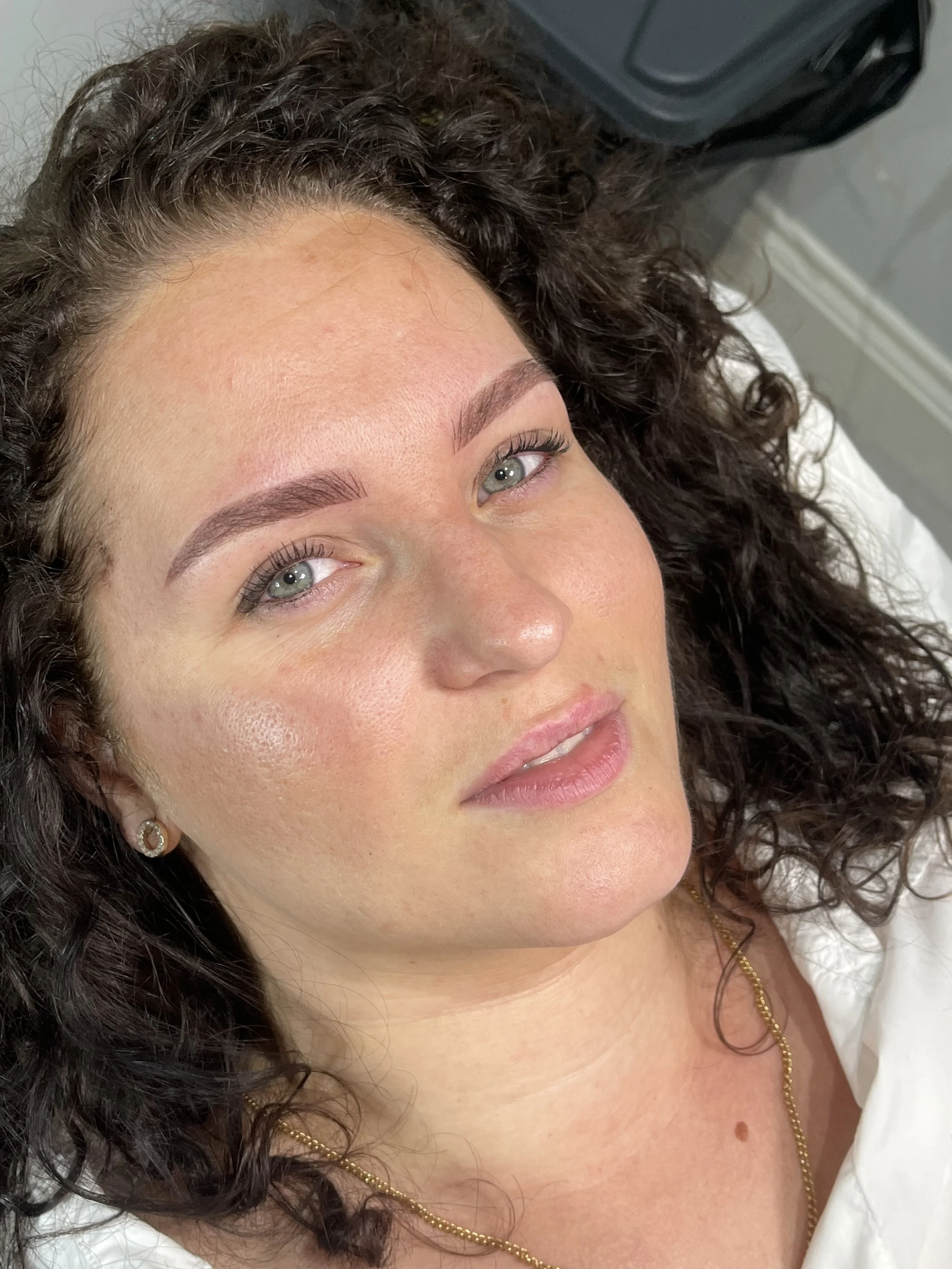 A woman with dark, curly hair lying down, looking at the camera with a slight smile, wearing earrings and a gold necklace.