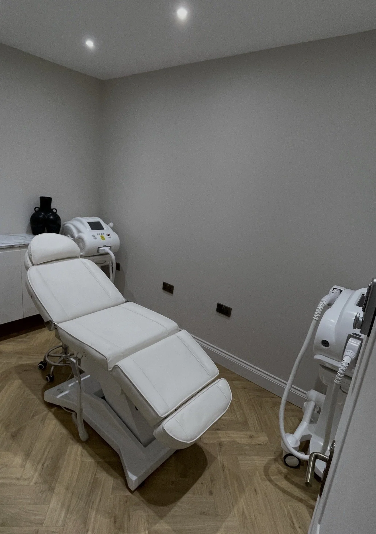 A treatment room with a white adjustable medical chair, two machines on either side, a white cabinet to the left, and a small black vase on top. The room has minimalist decor with beige walls and wooden flooring.