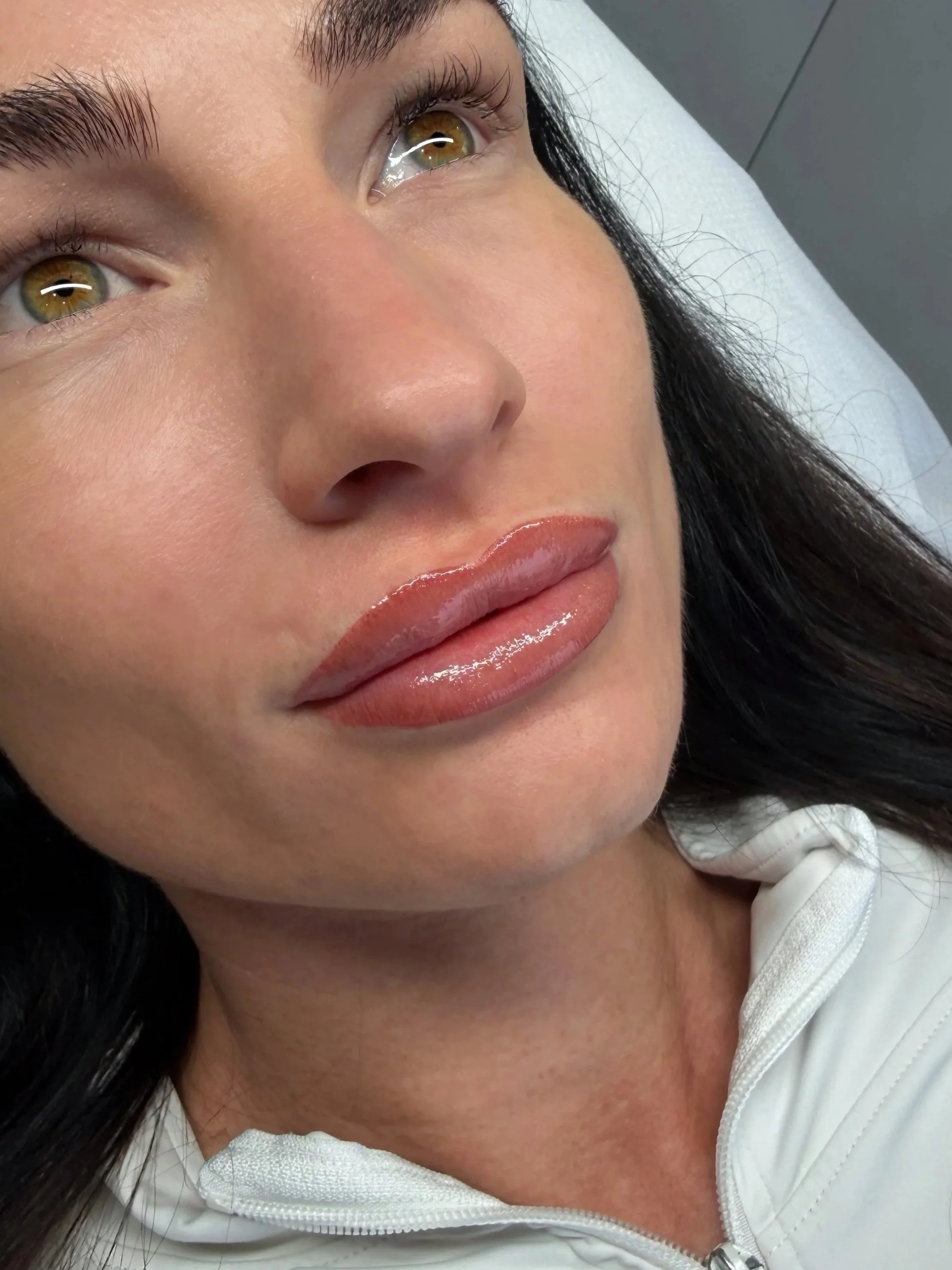 Close-up of a woman's face showing her hazel eyes, full lips with glossy lipstick, and dark hair, lying on a bed or examination table with a white blanket or sheet.