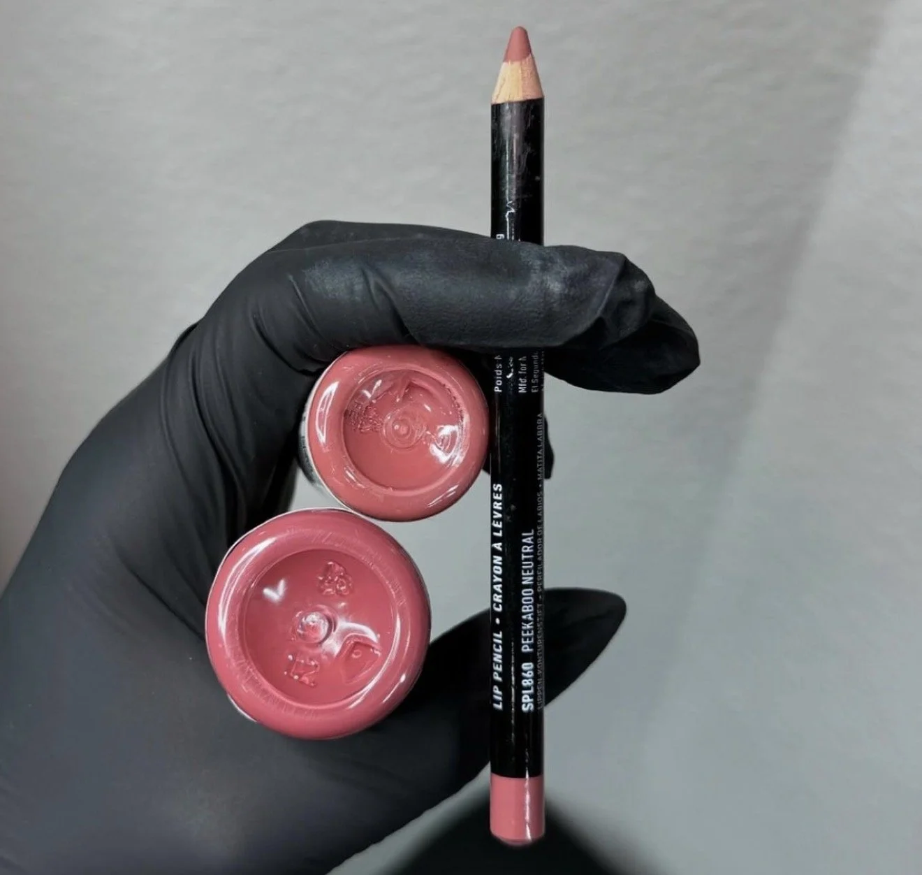 A hand wearing a black glove holding a pink lip pencil and two pink lipstick balm containers against a plain background.