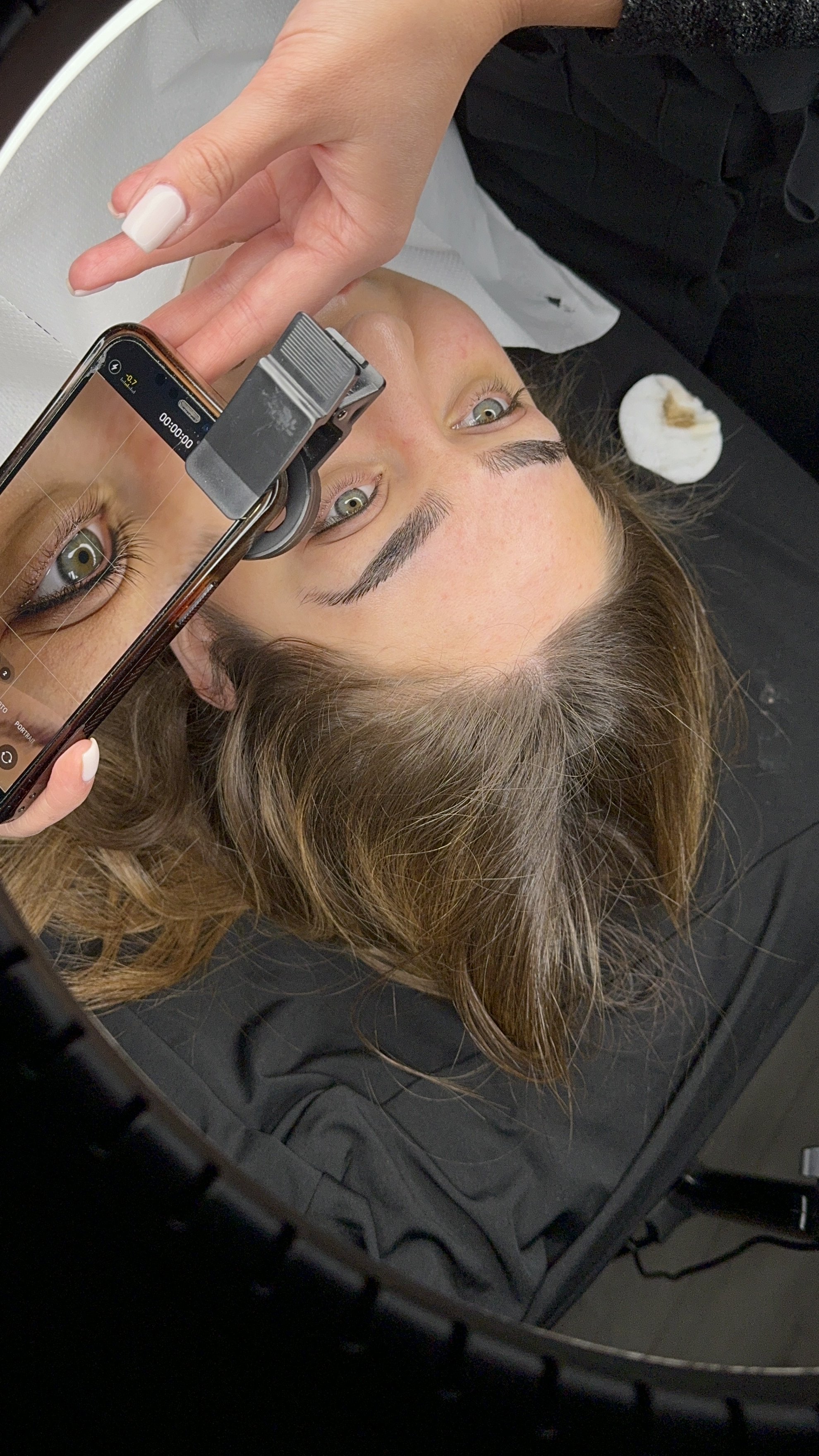 A woman lying on a black surface, taking pictures of her new eyeliner tattoo, using a small magnifying device,