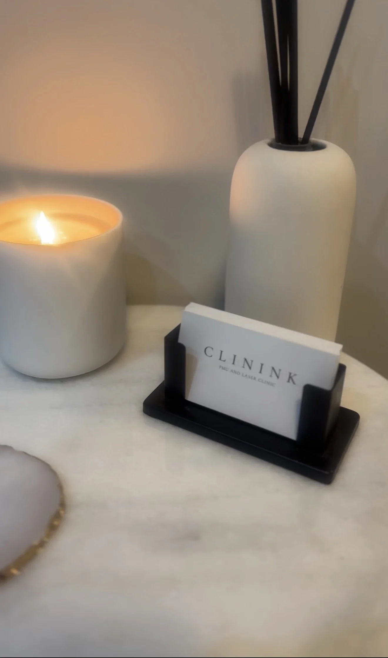 A candle, a business card holder with a card that says Clinink, a white vase with black sticks, and a small white plate with a gold rim on a white surface.