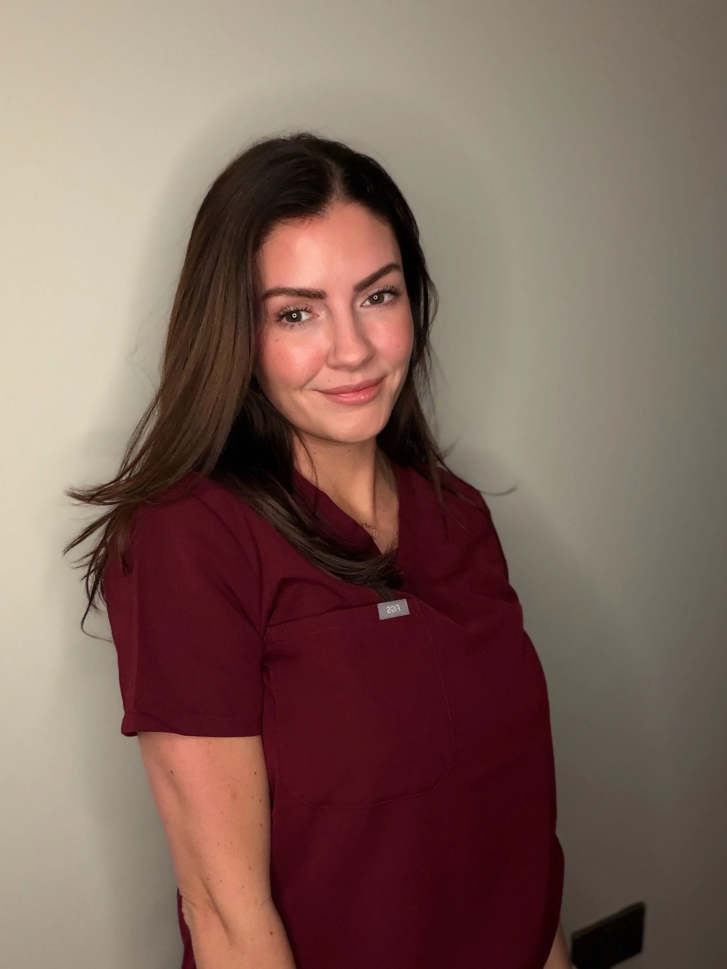 A woman with long brown hair wearing a dark red medical uniform, standing against a plain light-colored wall.