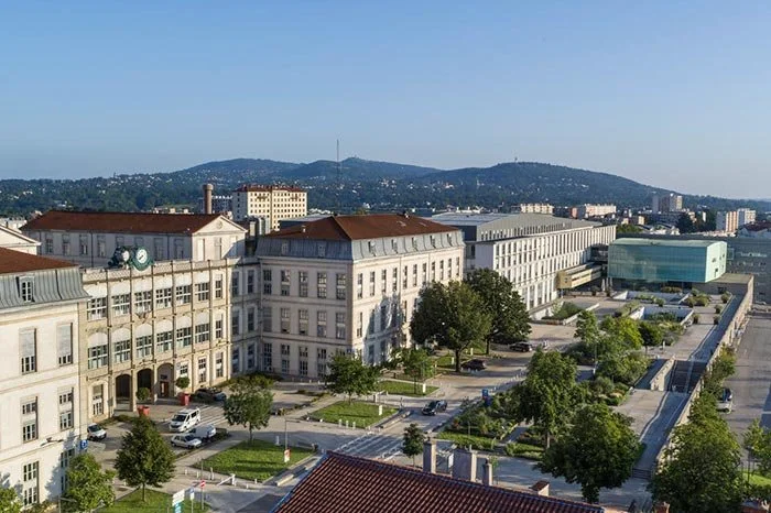 Vue aérienne d'un centre-ville avec des bâtiments historiques, des arbres et une montagne en arrière-plan, sous un ciel clair.