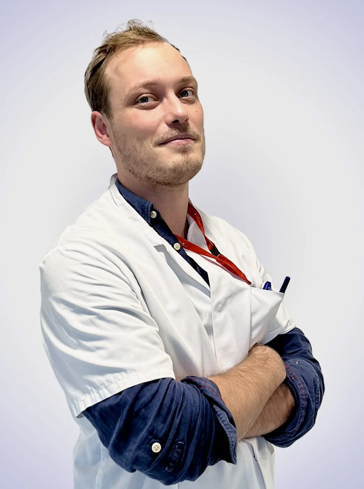 Un homme portant un costume médical blanc avec un badge rouge, croisé les bras, posant devant un mur blanc.
