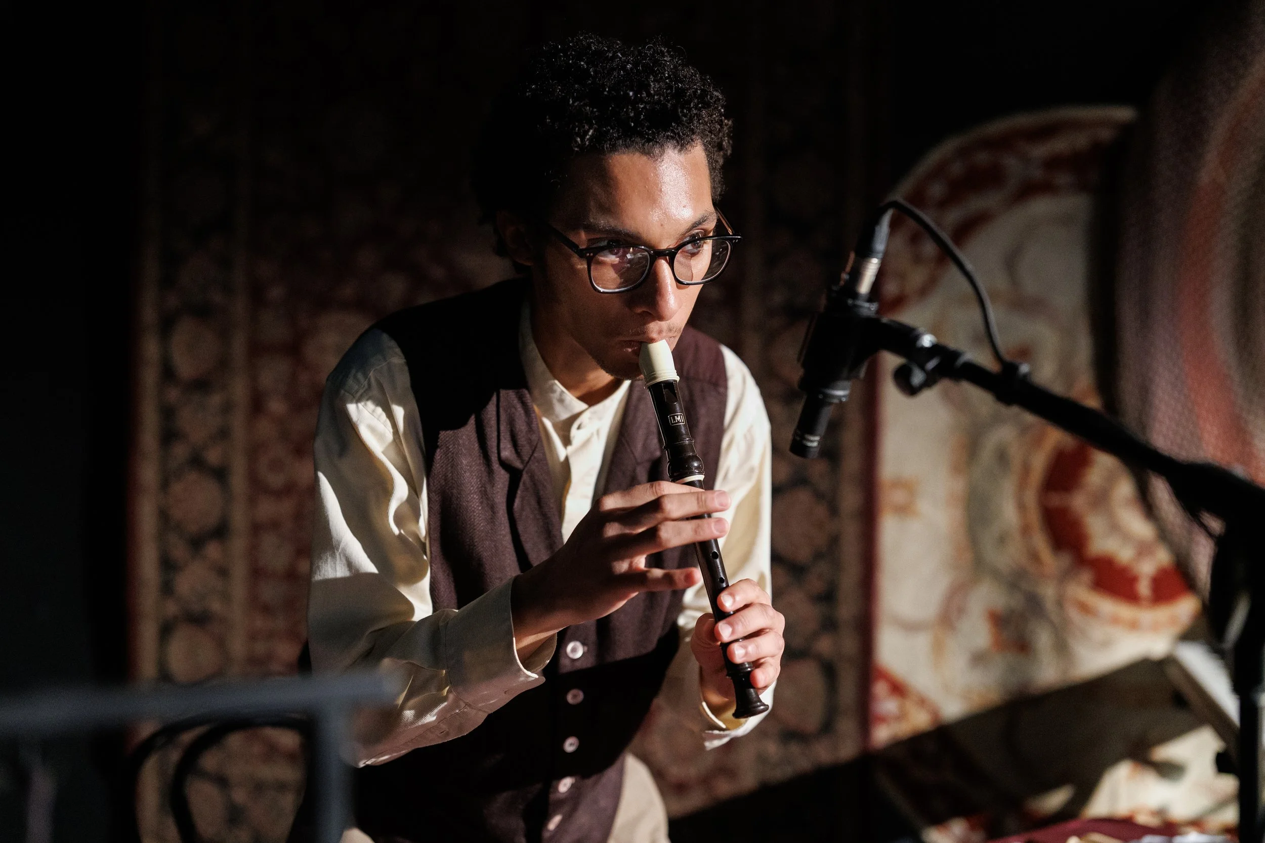 A person playing a recorder in a dimly lit room with a patterned rug on the wall in the background.