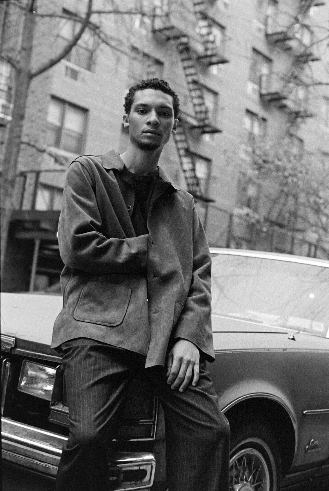 A young man with curly hair standing in front of a vintage car on a city street, with apartment buildings and fire escapes in the background, captured in black and white.
