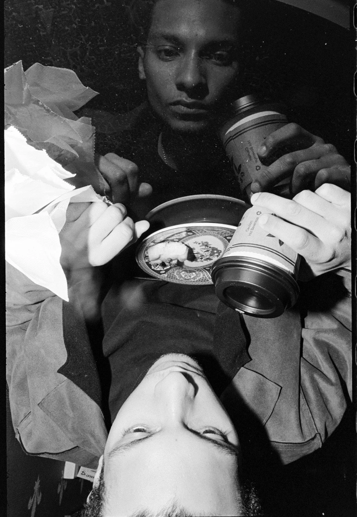 Two young men sitting at a table, each holding a paper coffee cup, with some torn paper and a plate with food on the table. The photo is in black and white, one man is facing the camera with their eyes closed, and the other is looking into the camera