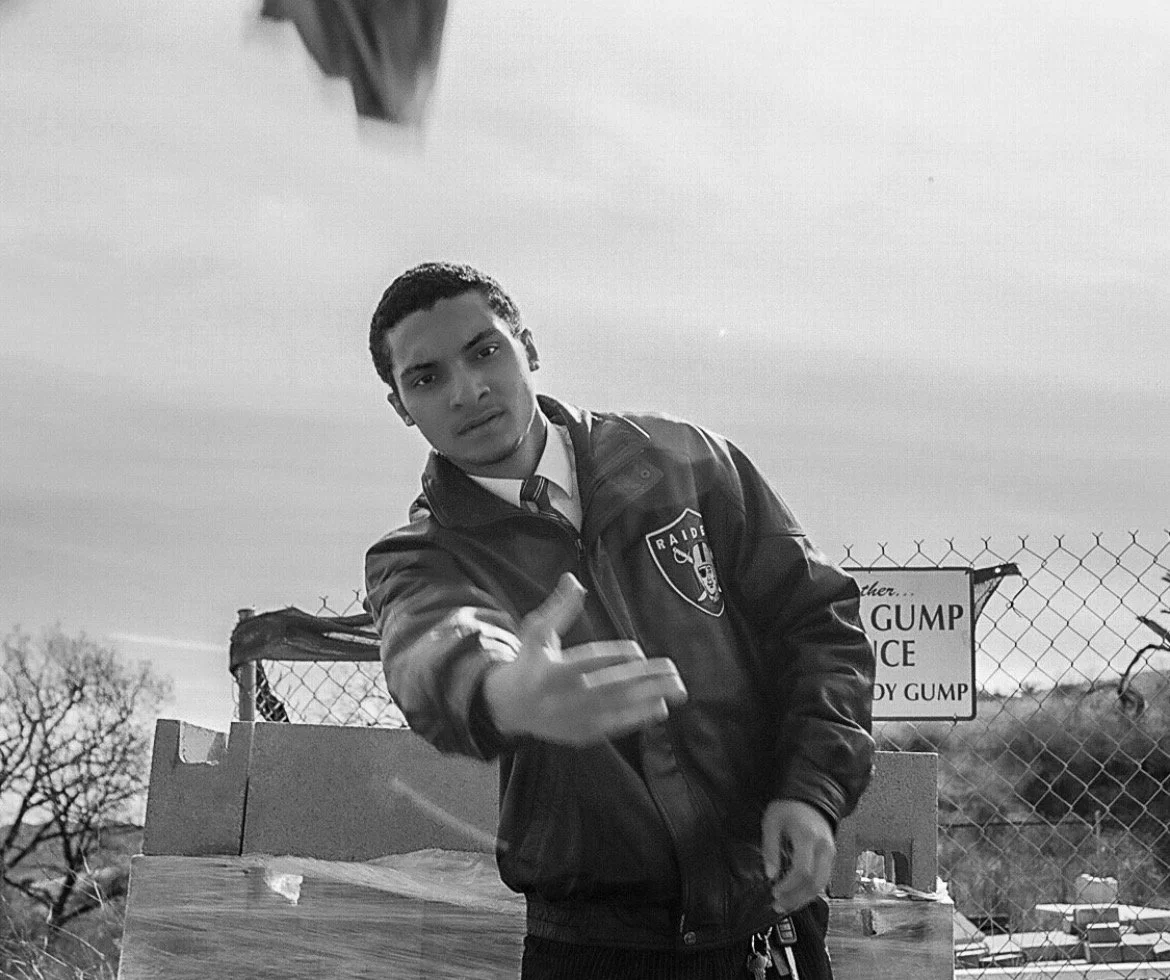 A young man in a Raiders jacket and tie making a hand gesture towards the camera outdoors next to a chain-link fence with a sign in the background.
