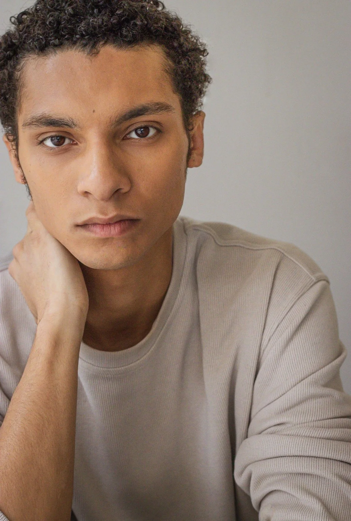 A young man with curly hair and brown eyes, wearing a beige shirt, looking directly at the camera with a neutral expression, resting his hand on his neck.