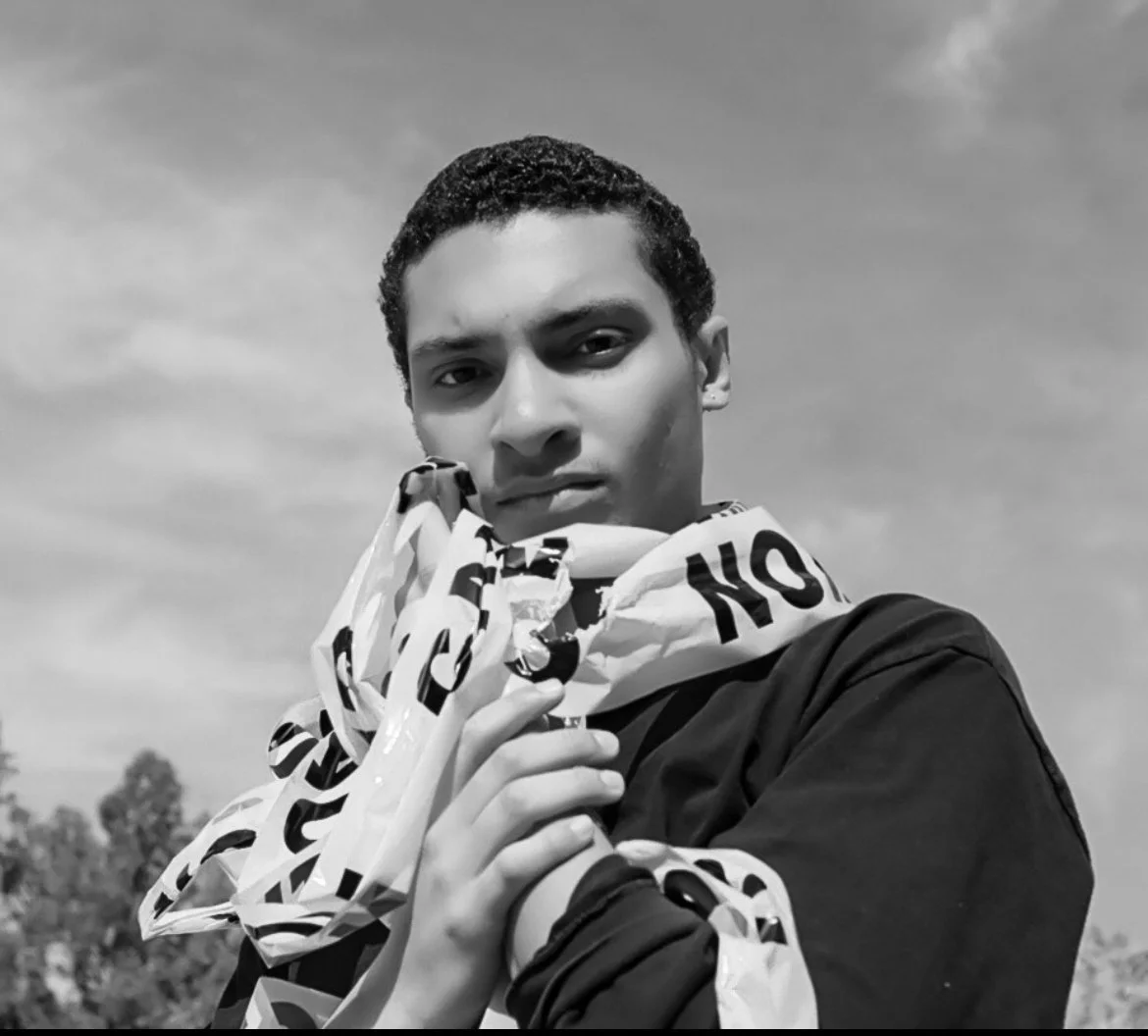 Black and white photo of a young person with short curly hair holding a crumpled patterned paper or plastic bag close to their chest, outdoors with a cloudy sky and trees in the background.