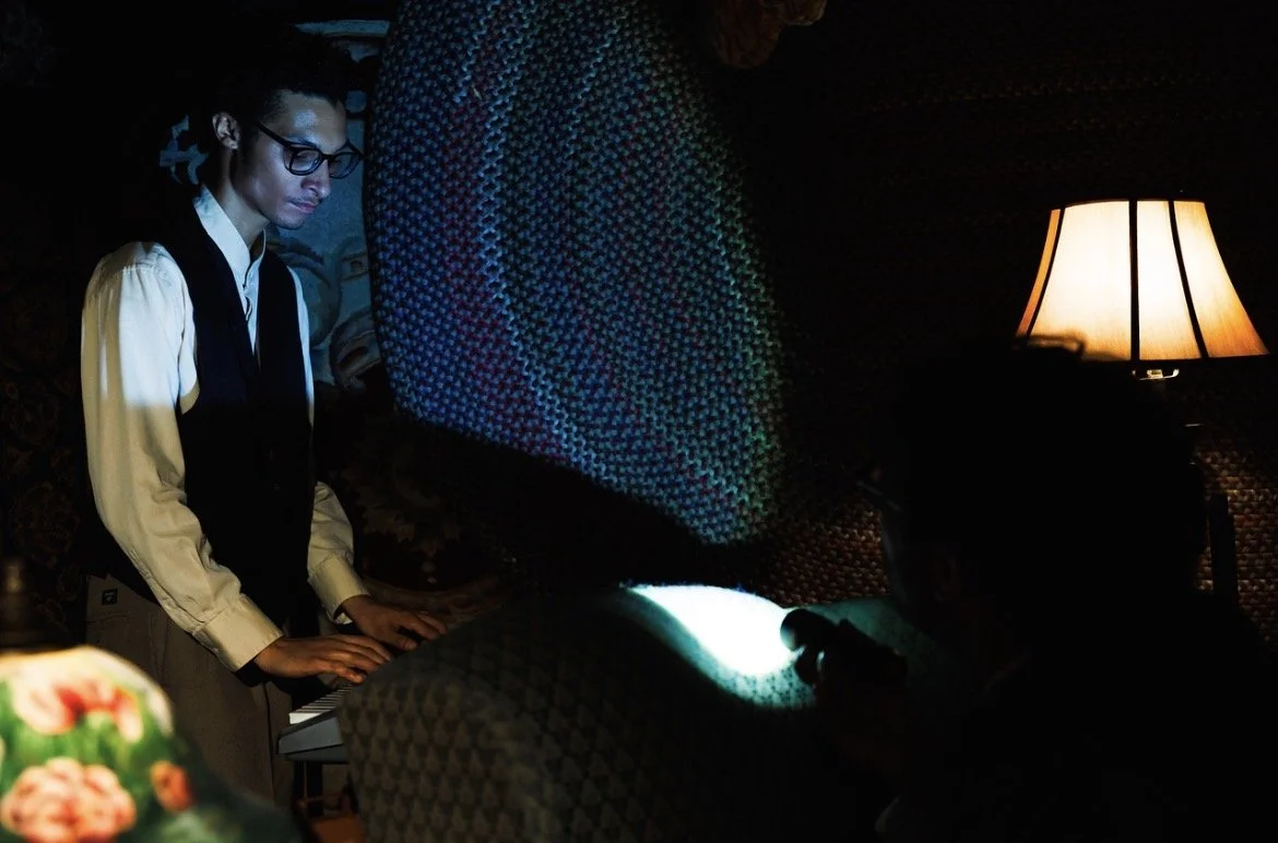 A woman playing the piano in a dimly lit room with a lamp on the side. She is concentrating on playing, wearing glasses and a formal outfit.