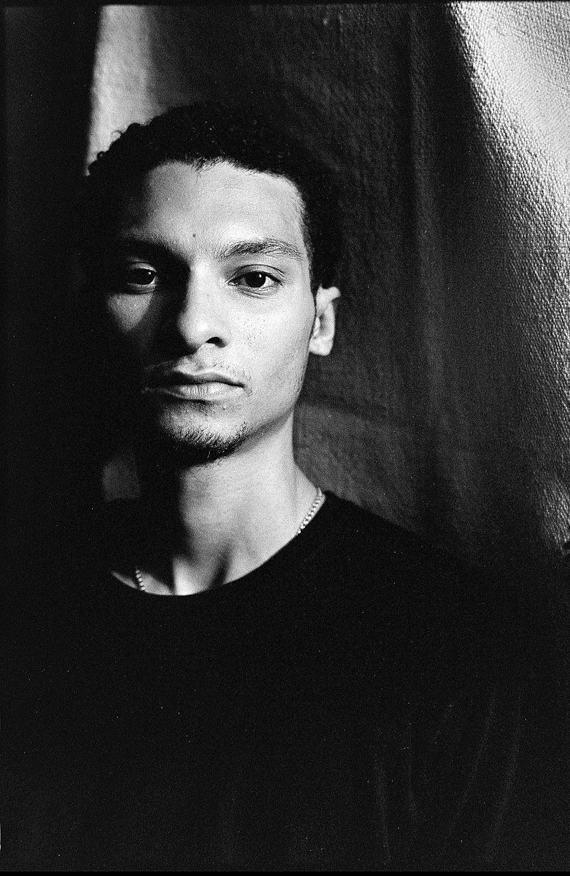 Black and white portrait of a young man with medium-length curly hair, wearing a dark shirt and a chain necklace, against a textured background.