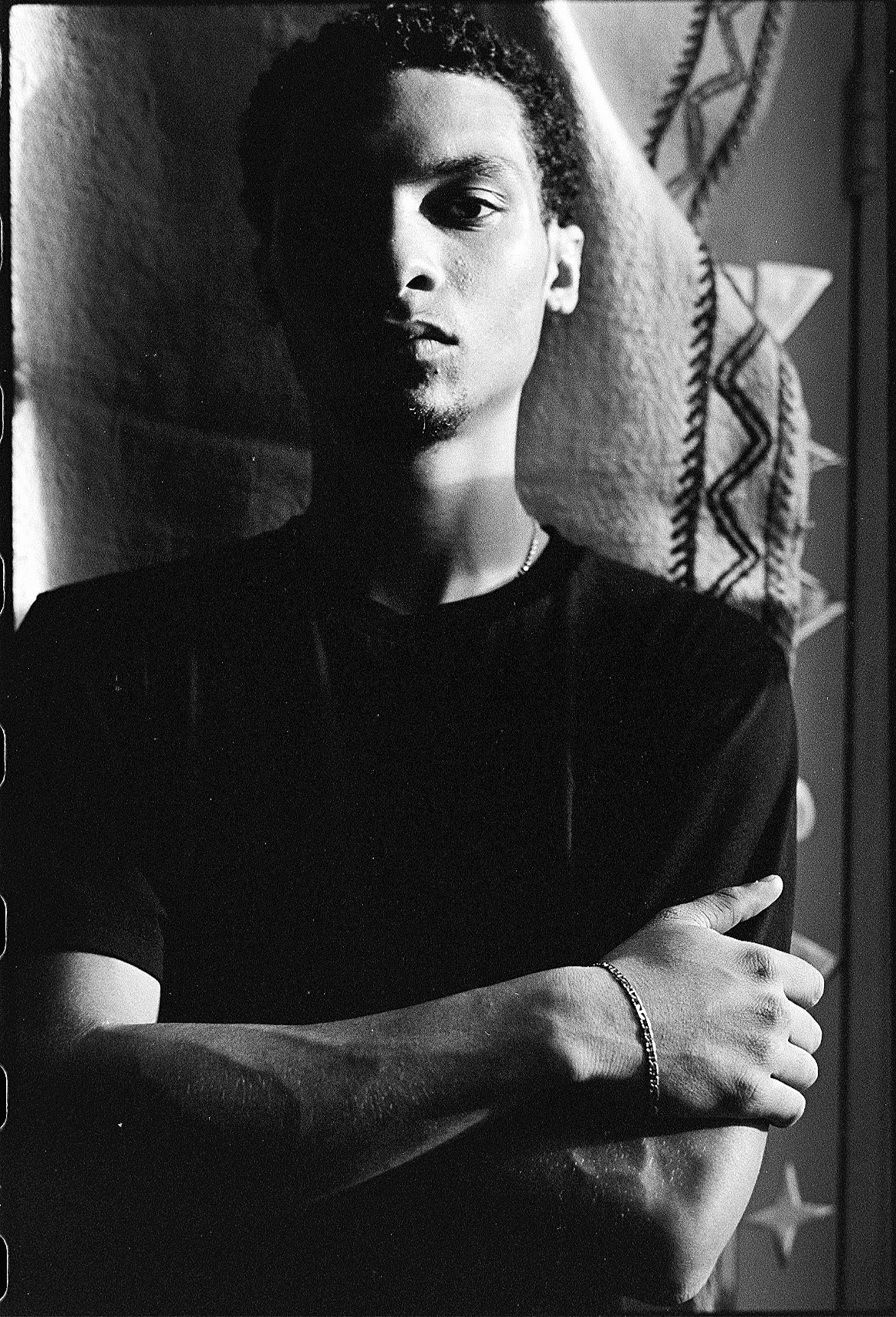 Black and white portrait of a young person with short hair, wearing a black t-shirt, standing with their eyes partially in shadow, their arms crossed, with decorative fabric or wall hanging in the background.