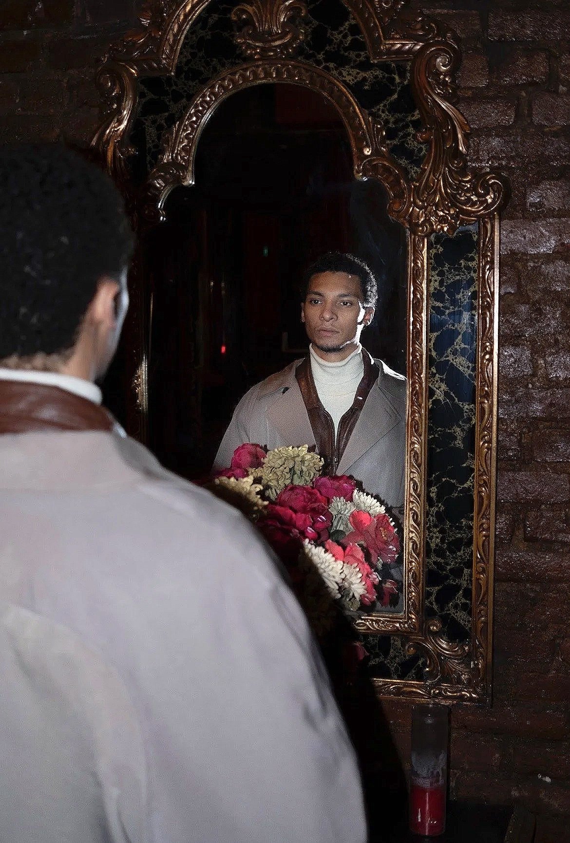 A man looking at his reflection in a vintage mirror, holding a bouquet of red, white, and pink flowers. The mirror has an ornate gold frame and the background features a brick wall.