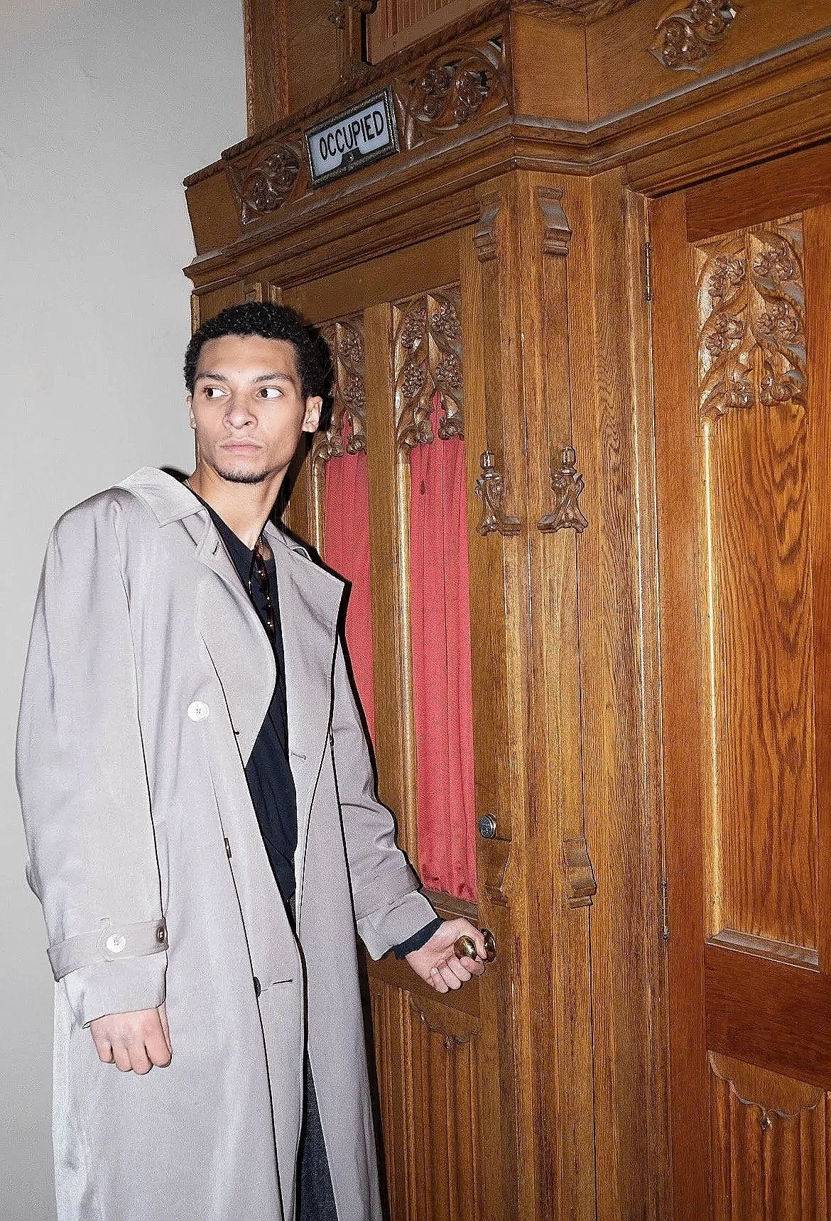 A young man with dark, styled hair wearing a beige trench coat over a black shirt is standing in front of a wooden door. He is slightly turned to the side, holding the door handle with his right hand, and looking to his left. The door has intricate w