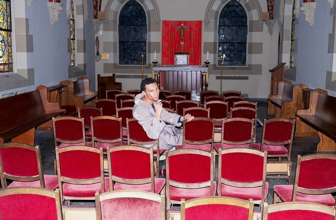 A young man sitting in an empty church with red velvet chairs, stained glass windows, and religious symbols on the altar