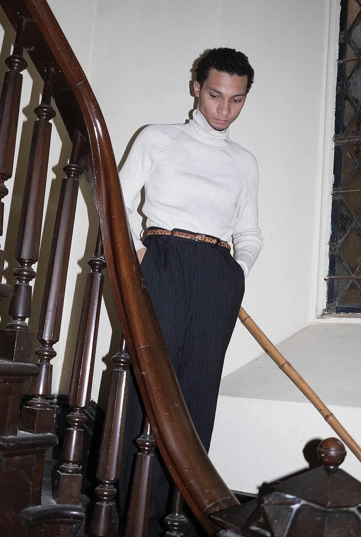 A young man with curly dark hair, wearing a white turtleneck sweater and dark striped trousers with a leopard-print belt, standing on a staircase with wooden banisters and holding a bamboo stick, looking down.