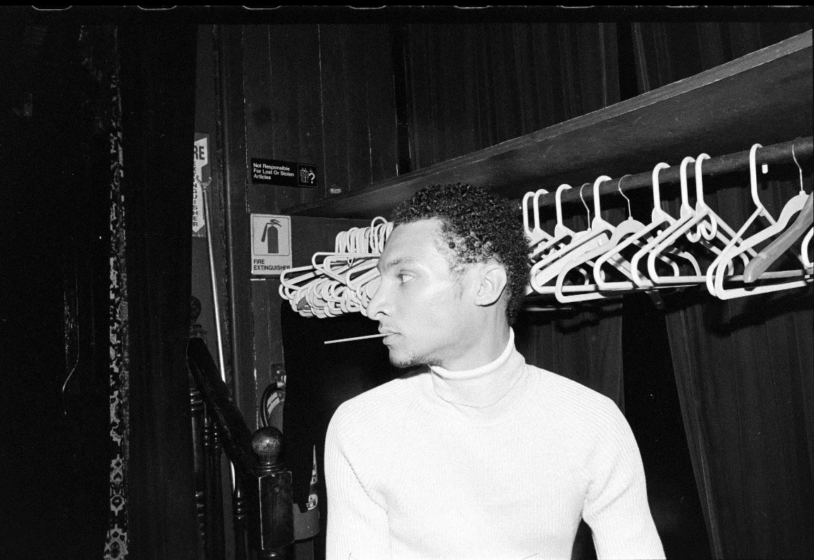A Black and white photo of a man with curly hair and a mustache, wearing a light-colored turtleneck, looking to the left in front of a rack of empty hangers in a dark room.