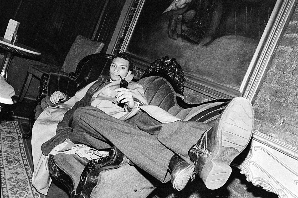 A man sitting on a vintage sofa in a room with dark wood paneling, holding a pipe, with a large framed mirror or painting behind him.