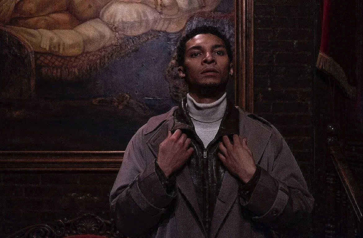 A young man with dark curly hair stands in front of a dark wall with a framed painting behind him. He is wearing a light gray jacket over a black shirt and a white turtleneck, holding the collar of his jacket with both hands, looking directly at the 
