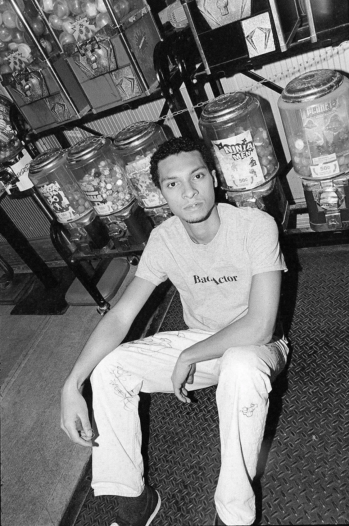 A young man with short curly hair and facial hair sitting on a bench in front of a row of gumball machines in an arcade or amusement center. He is wearing a t-shirt and light-colored pants.
