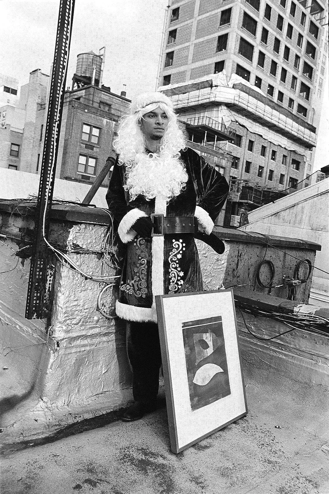 A person dressed in a Santa Claus costume with a wig, standing on a rooftop in an urban area, with a framed abstract artwork leaning against the ledge and a cityscape of high-rise buildings in the background.