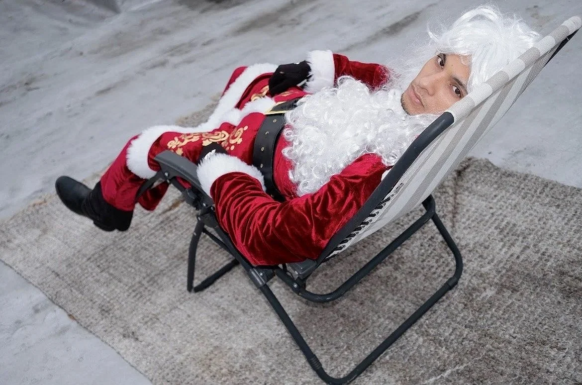 A person dressed as Santa Claus lying on a lounge chair on a beige rug, indoors with a gray marble floor. They are wearing a red velvet Santa suit with white trim, black gloves, black boots, and a curly white beard and wig. They appear relaxed and ar