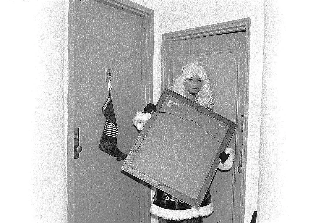 Person dressed in Christmas costume holding a framed picture, standing in front of a door with a holiday sock hanging on the door handle.