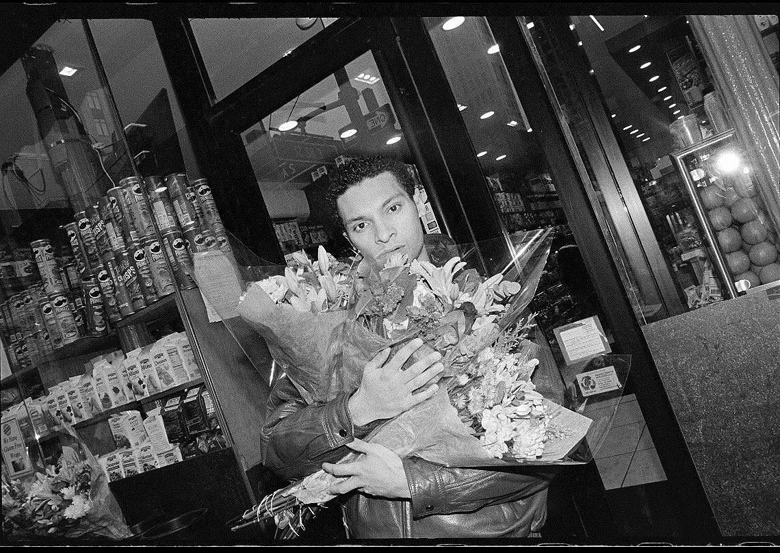 A young man holding a large bouquet of flowers in front of a store window, with grocery items and a gumball machine visible behind him.