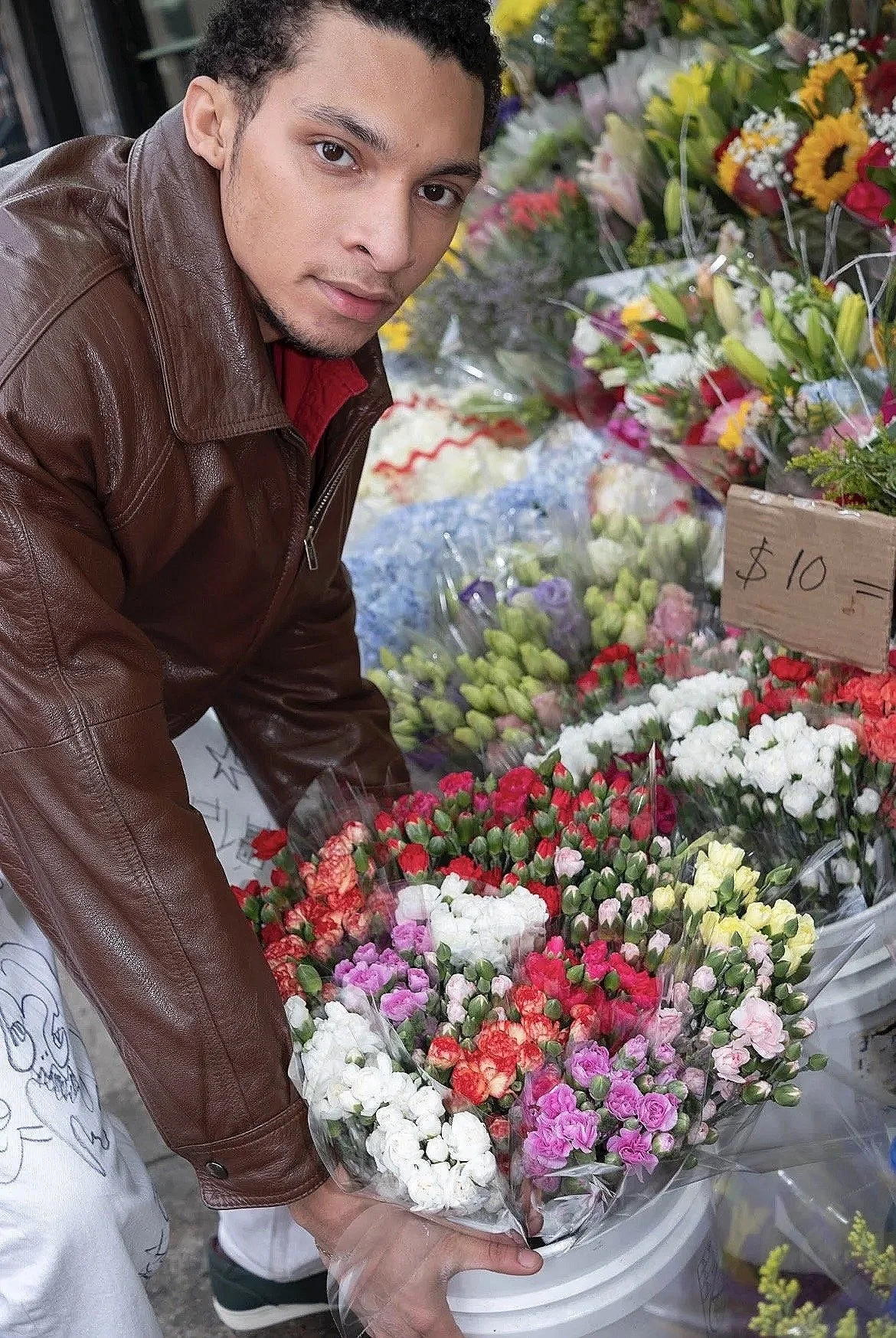 A young man wearing a brown leather jacket leans over a display of colorful flowers at a flower market, holding a bouquet of pink, white, and red blooms wrapped in plastic. A handwritten sign indicates the price of $10.