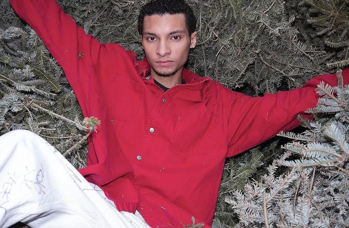 A young man with dark hair and a mustache lying among pine branches, wearing a red jacket and white pants.