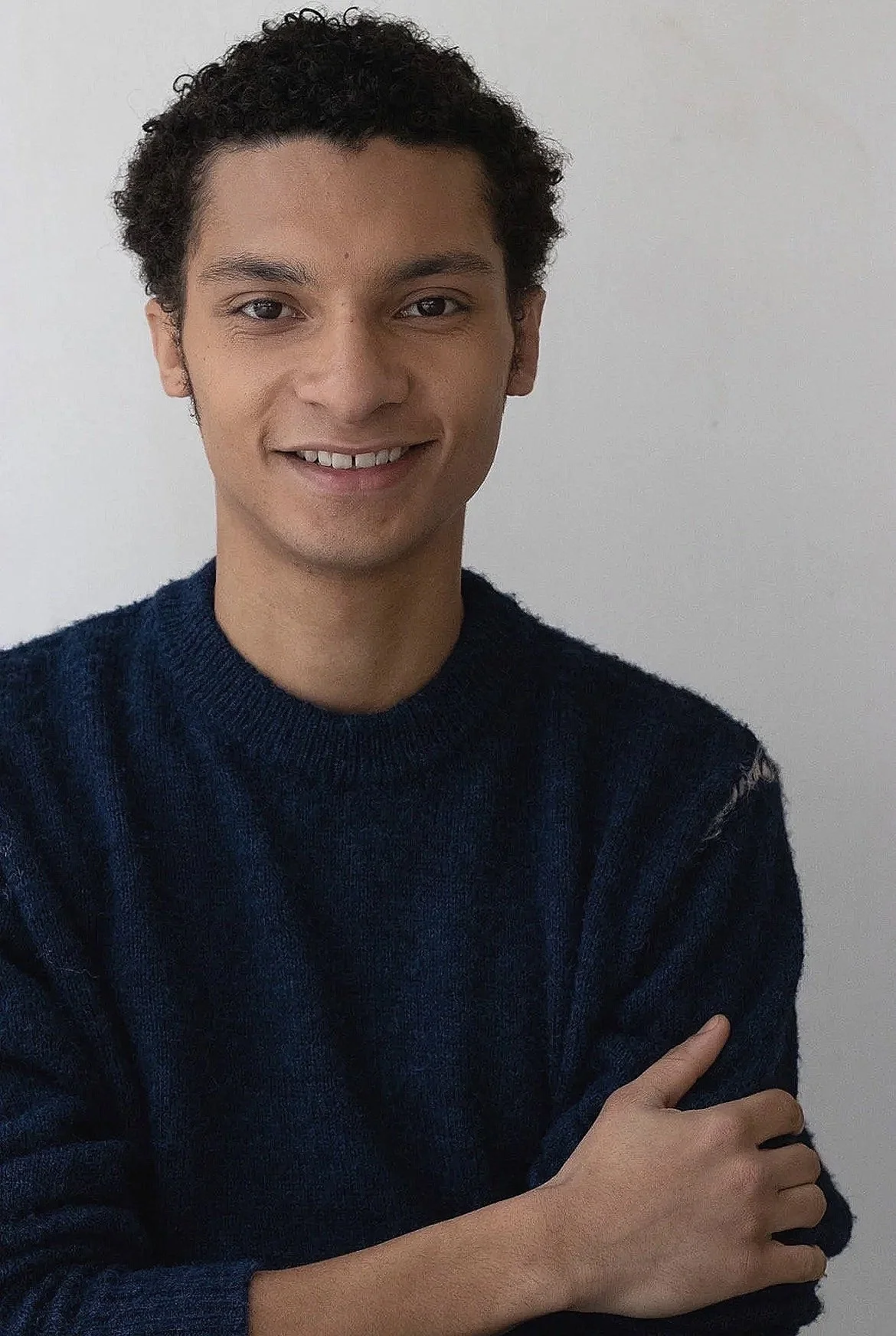 Young man with short curly dark hair, smiling, wearing a navy blue sweater, standing against a plain white background.