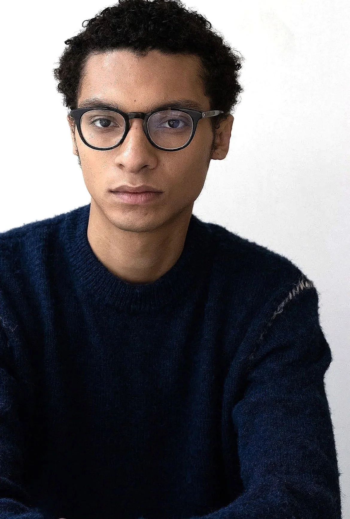 A young man with curly black hair wearing glasses and a navy blue sweater, looking directly at the camera against a plain white background.