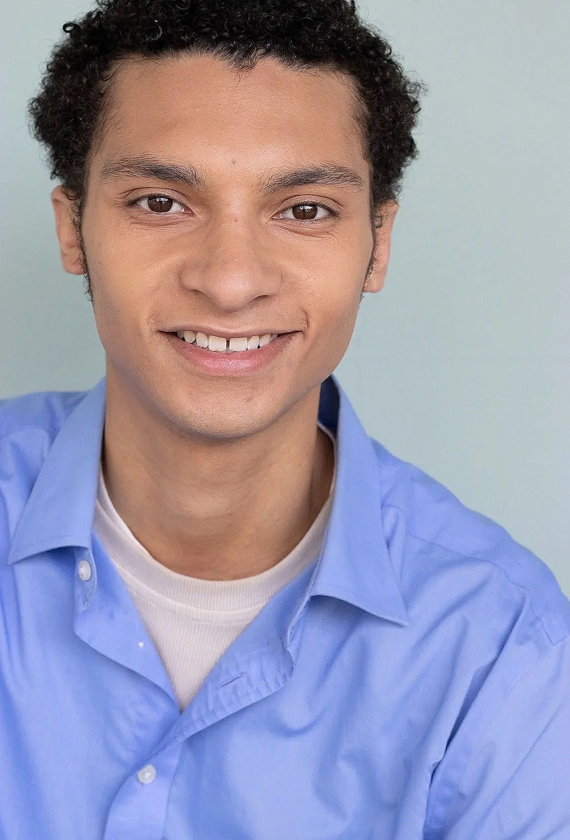 A young man with short curly black hair, brown eyes, and light brown skin, smiling while wearing a light blue button-up shirt over a white t-shirt, against a light background.