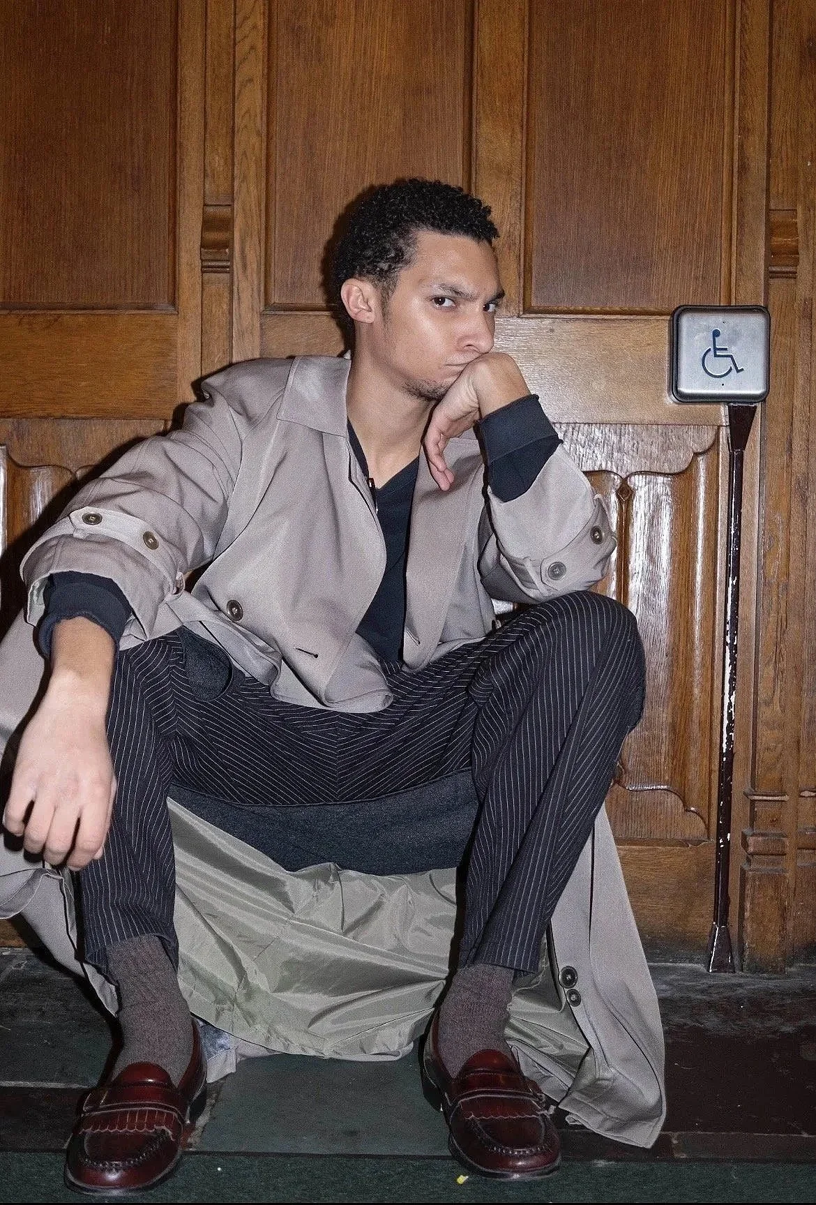 A young man sitting on the floor in front of a wooden wall, squatting with his hand on his chin, wearing a beige jacket, striped pants, and loafers, with a handicap parking sign in the background.