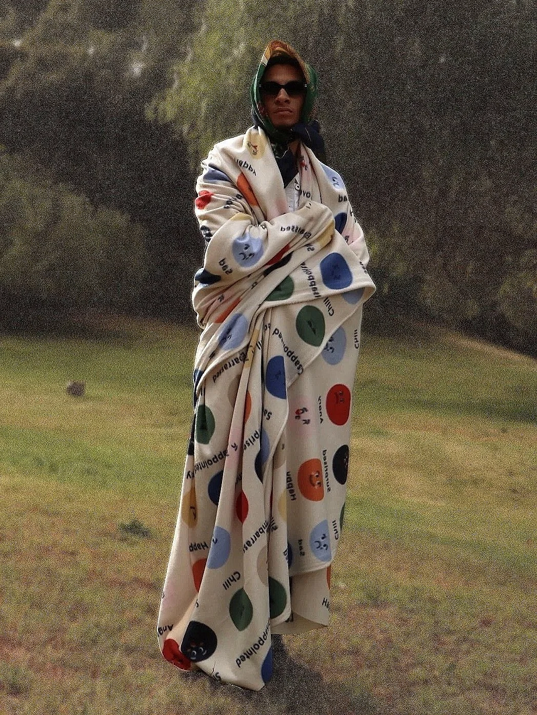 A person standing outdoors on a grassy field, wearing sunglasses, a helmet, and a massively oversized blanket with a colorful Polaroid pattern.