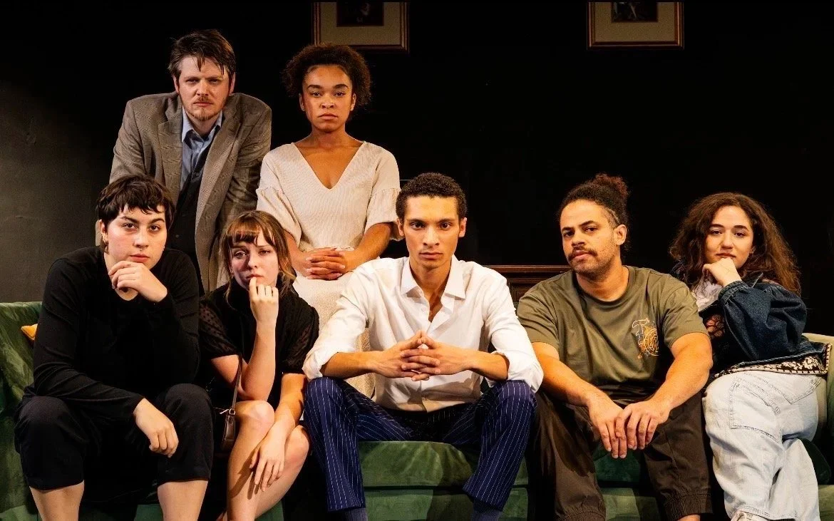 Group of seven young actors on stage in a drama play, sitting and standing with serious expressions.