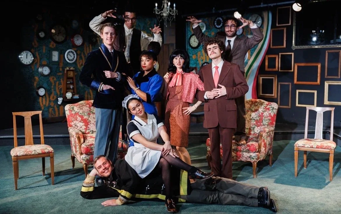 Seven actors in vintage costumes posing on a stage set resembling a living room, with a patterned wallpaper, clocks, and furniture, during a theatrical performance.