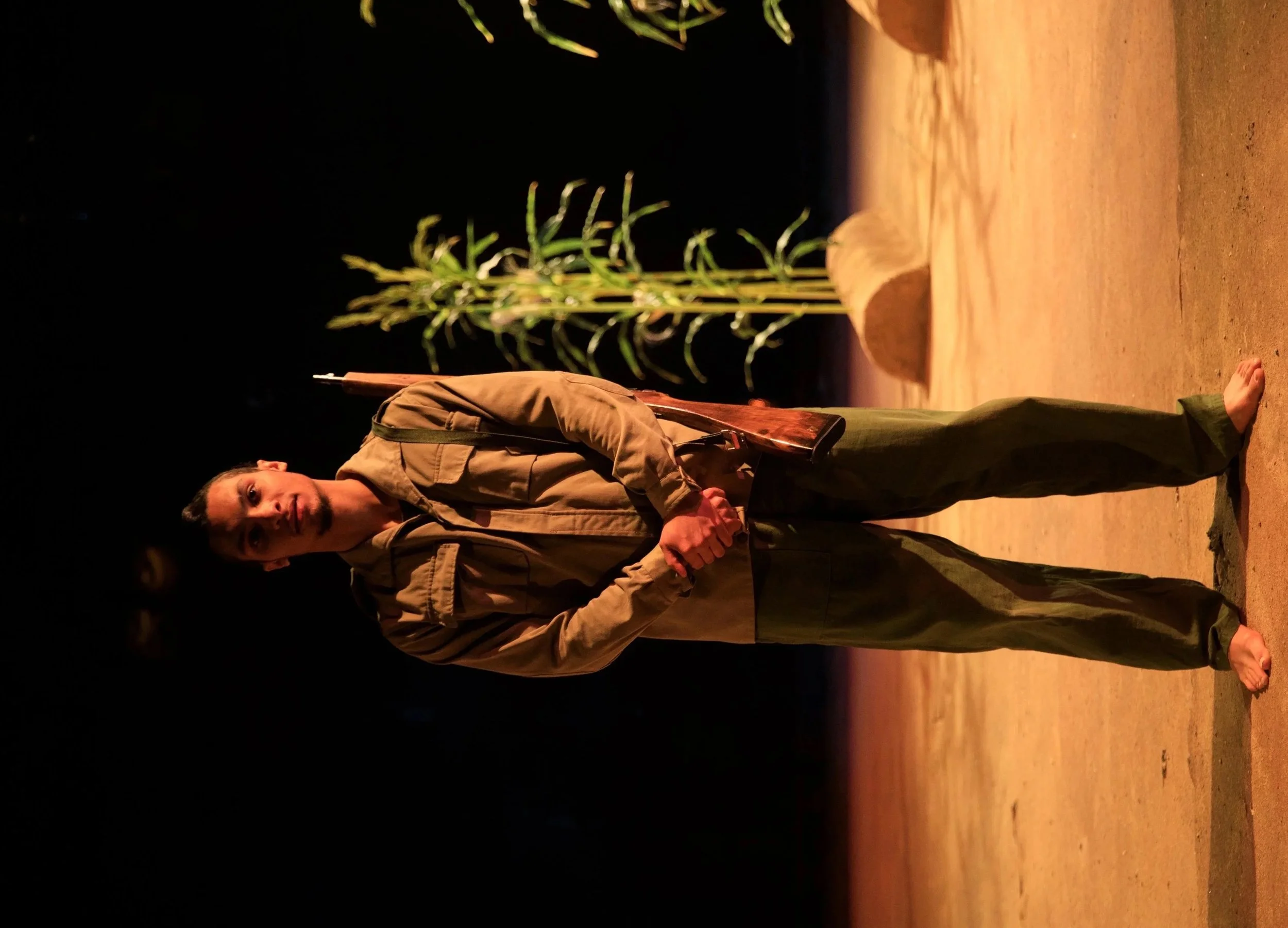 A man in military uniform holding a rifle standing barefoot on sandy ground at night with green crops and sandbags in the background.