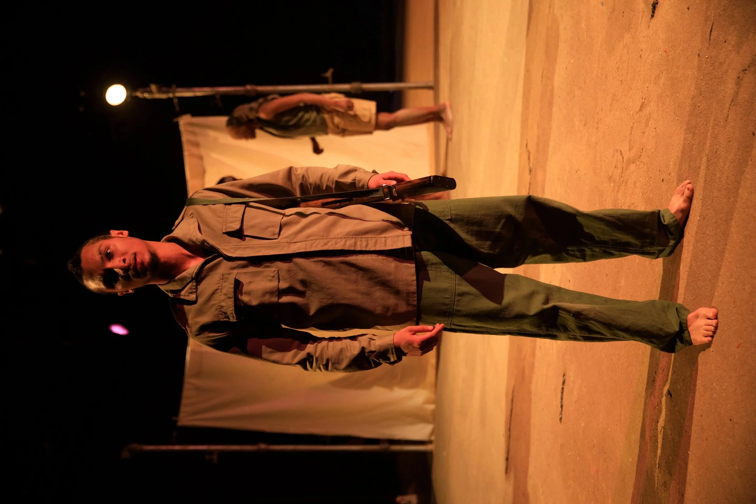 A man stands barefoot on a stage holding a microphone, with a woman and a white screen in the background.