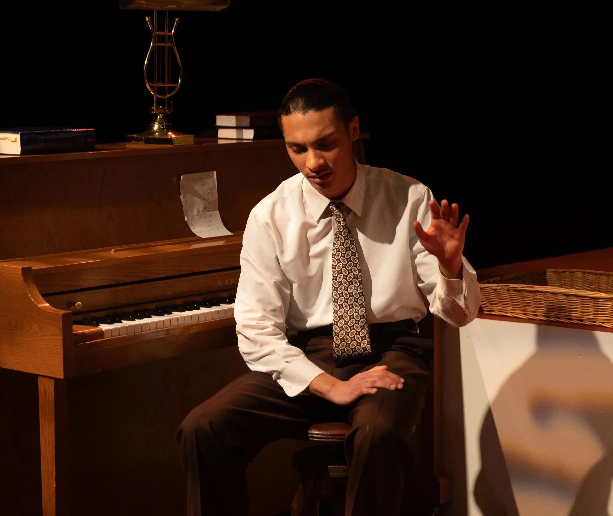 A man in a white shirt and patterned tie sitting on a stool next to a piano, gesturing with his right hand while speaking or singing, with a dark background and a few books and a decorative object on the piano.