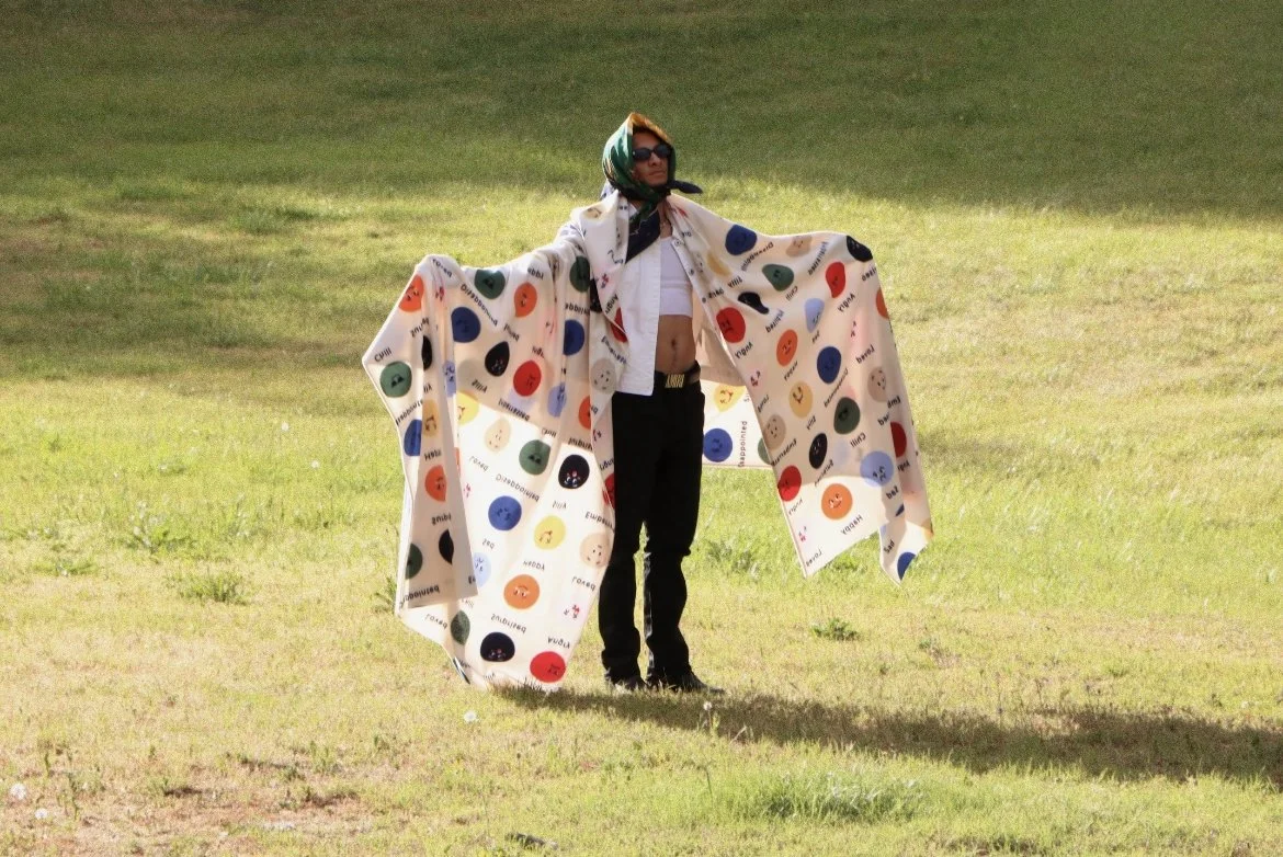 Person standing outdoors on grass, wearing sunglasses, a white shirt, black pants, a colorful scarf on their head, and a large cloth with multicolored dots and words draped over their arms.