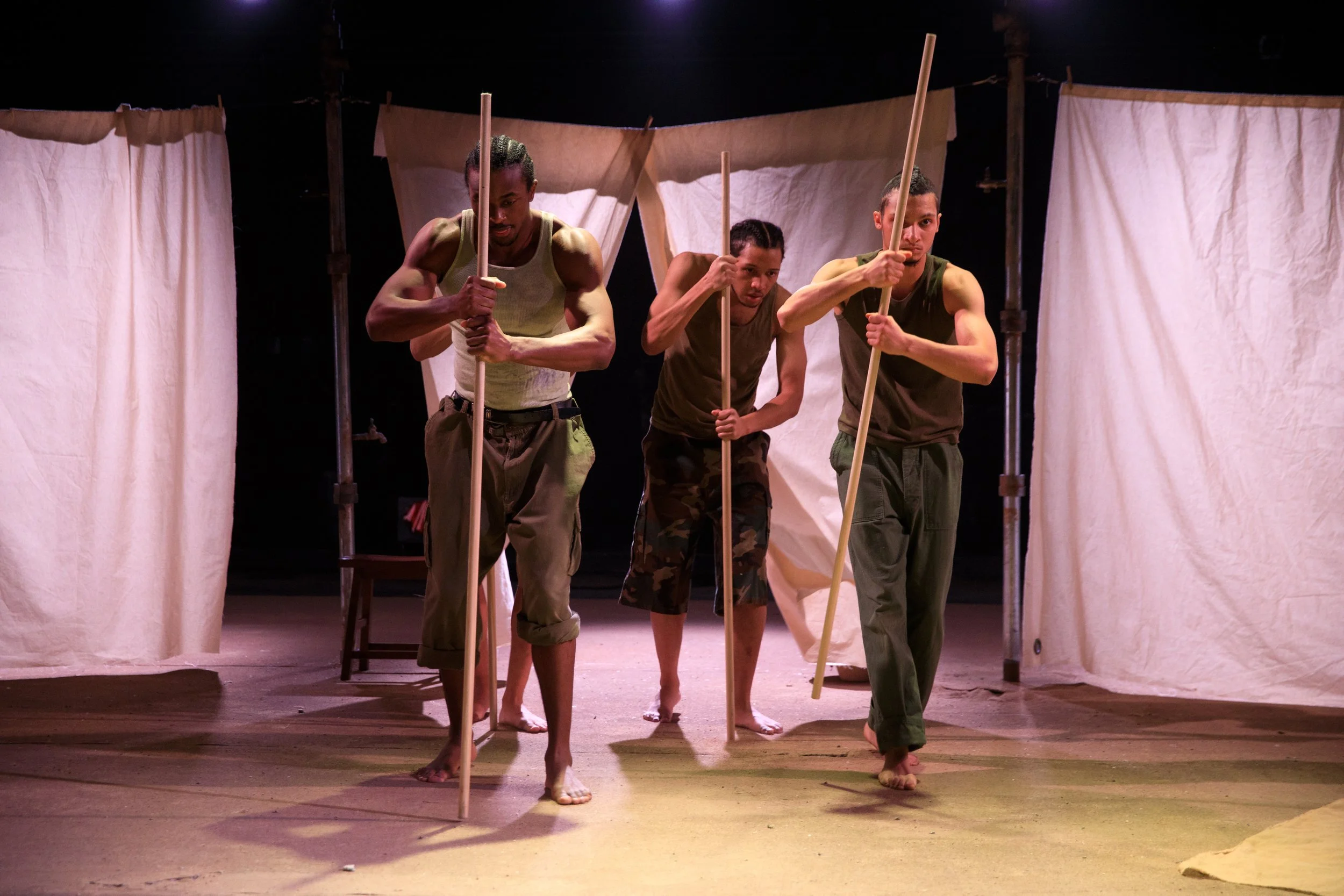 Three male performers on stage in a theatrical production, holding long sticks, with beige curtains in the background.