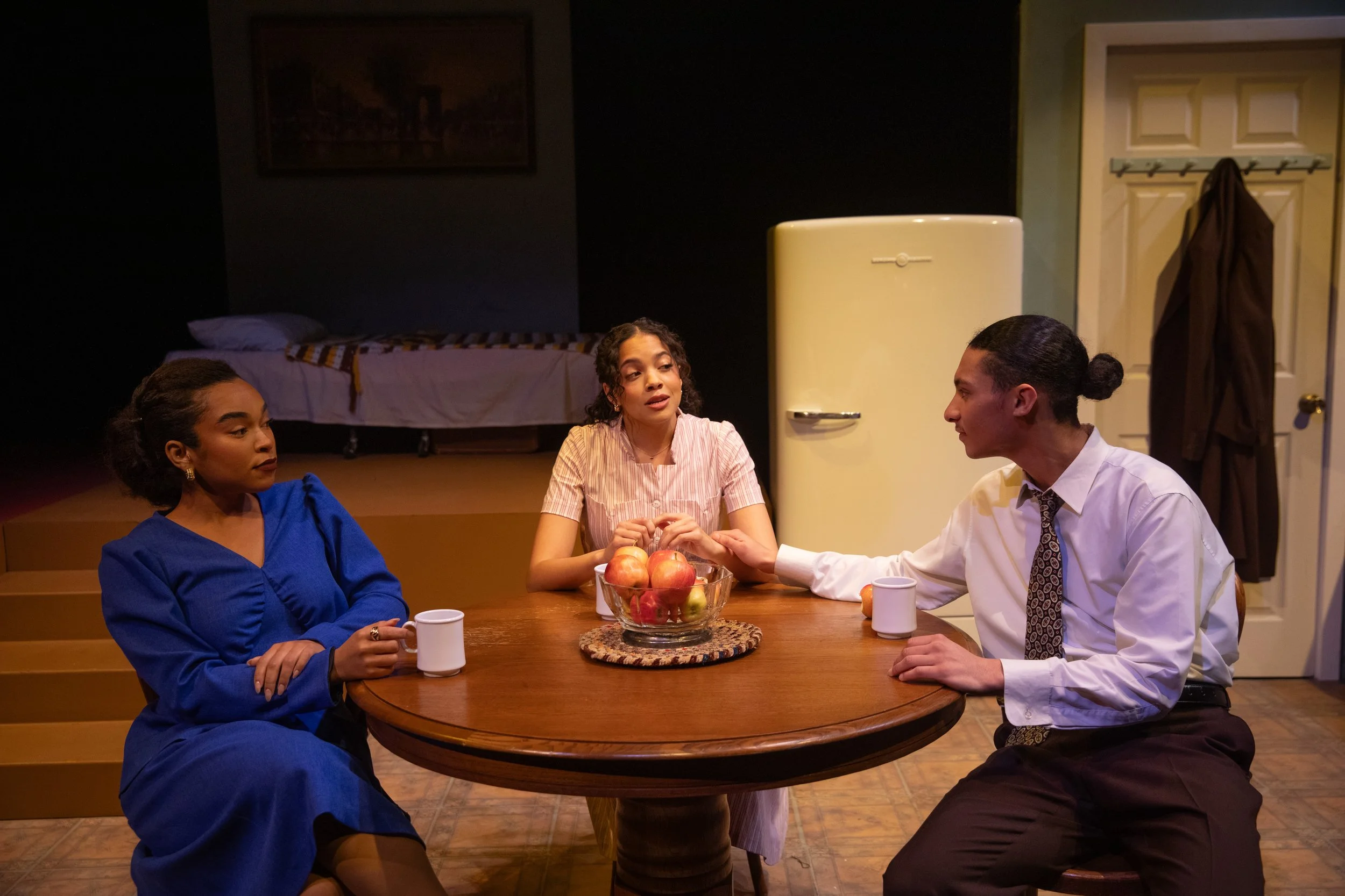 Three people sitting at a round wooden table having a conversation, with a bowl of apples in the center. The woman on the left is wearing a blue dress, the woman in the middle is wearing a pink striped shirt, and the man on the right is wearing a whi