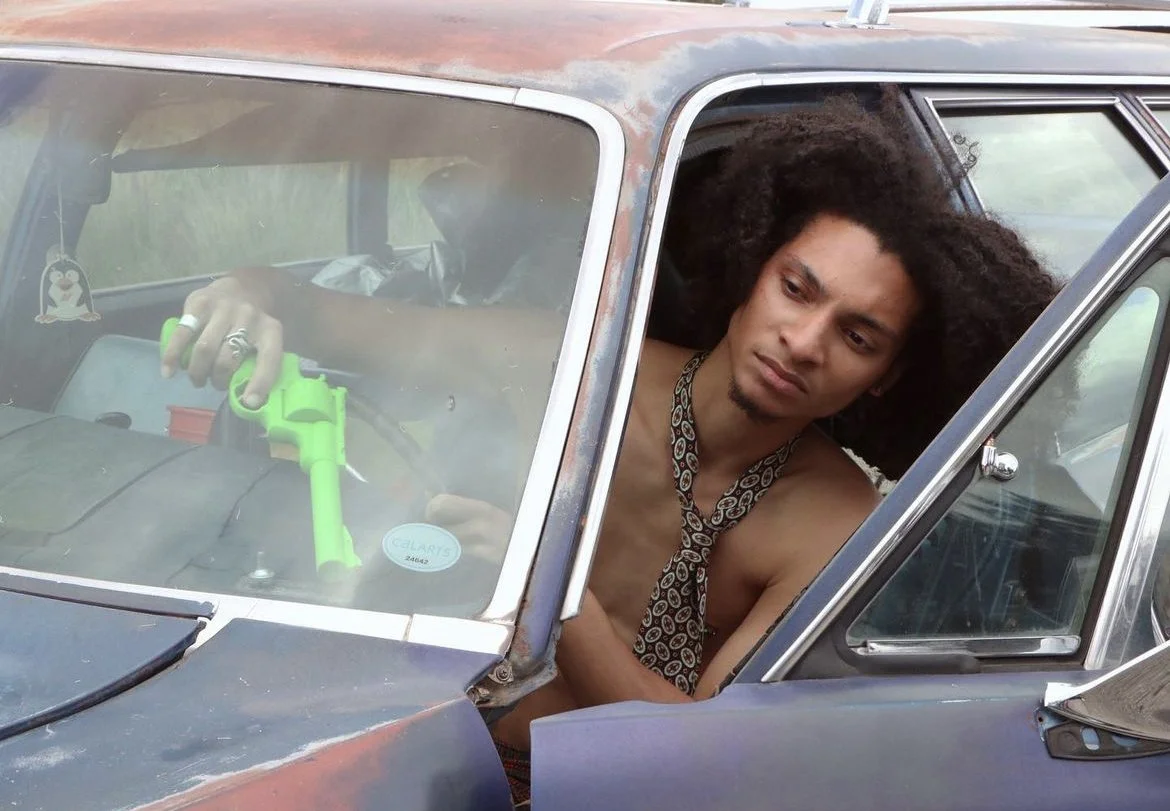 Young man with curly hair and a patterned tie around his neck sitting inside an old, rusty car, looking at a bright green toy gun he is holding in the dashboard area.