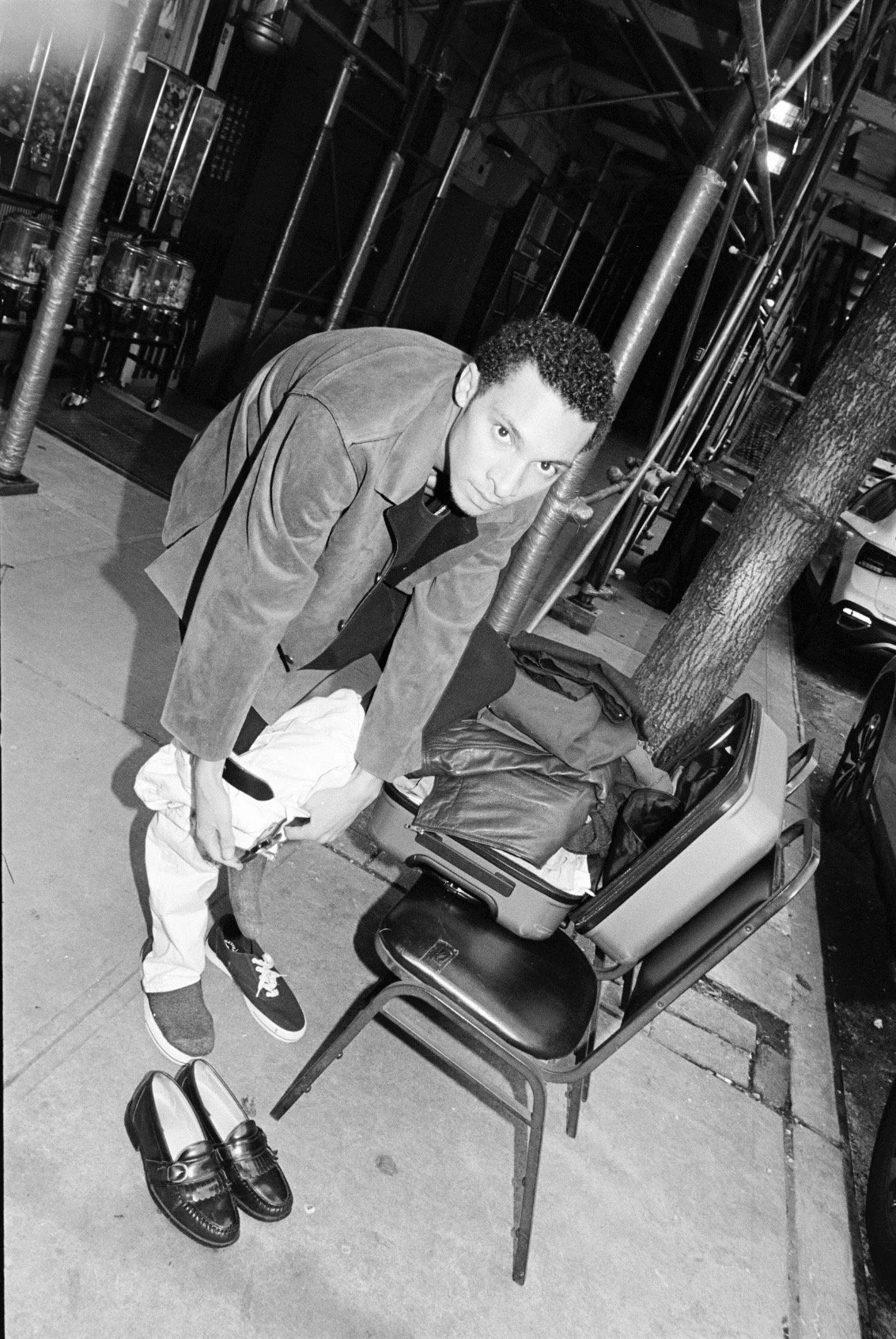A man in a jacket and sneakers kneels beside a chair on a city sidewalk at night, with a suitcase and bags filled with clothes nearby and a pair of loafers on the ground.