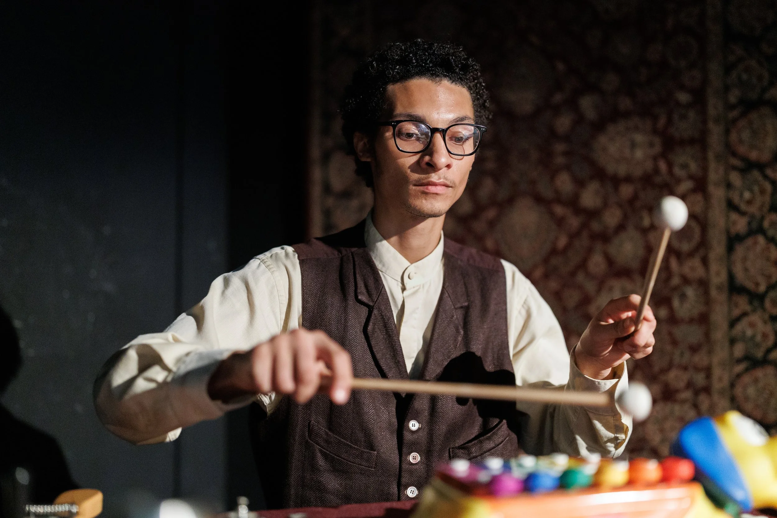 A man with glasses and curly hair playing a musical instrument with mallets, colorful objects, and a wire sculpture on a table in front of him.